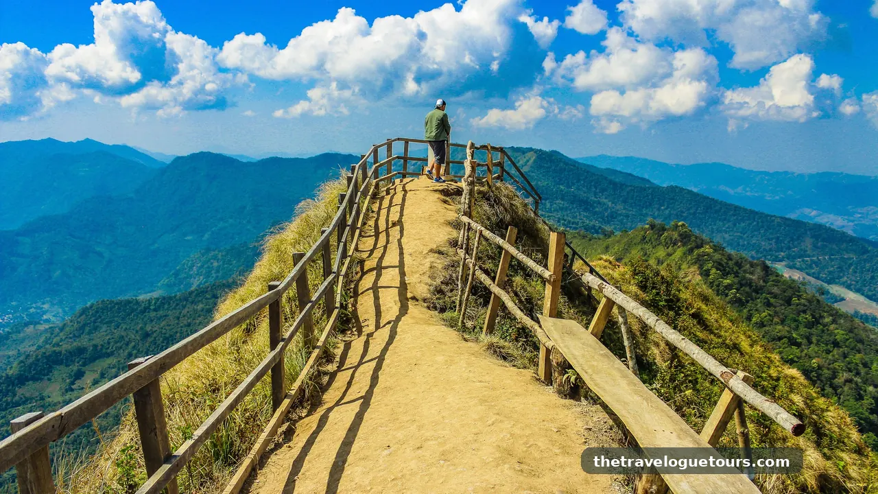 A hiker walking a scenic mountain trail surrounded by lush mountains and clouds